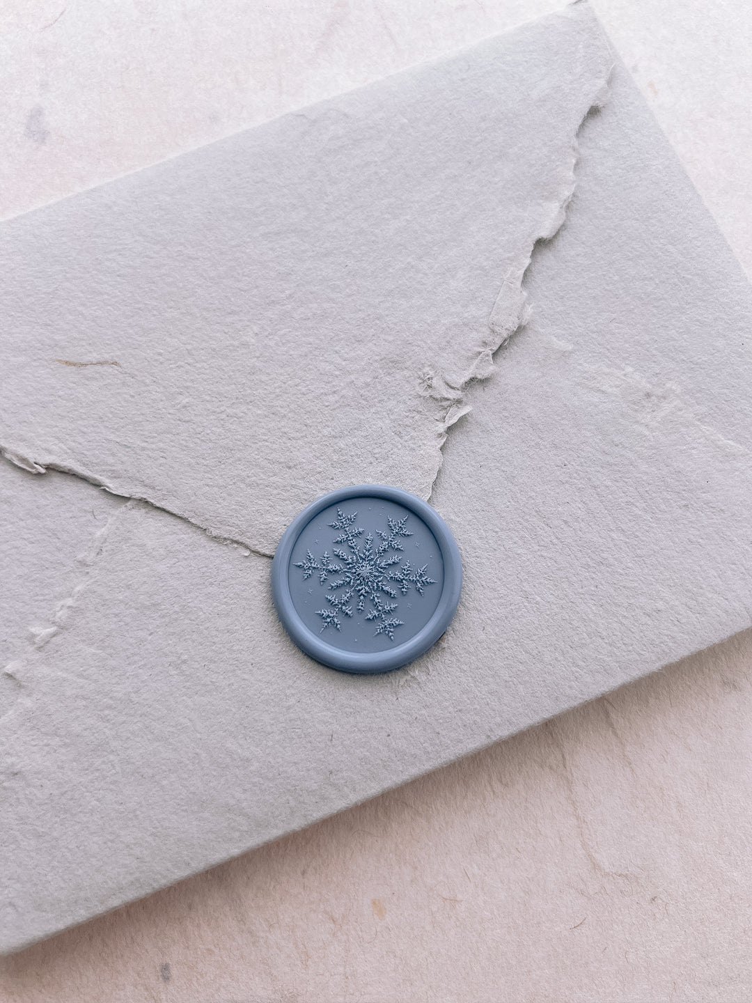 Dusty blue 3D engraved snowflake wax seal on a light grey deckled edge handmade paper envelope 