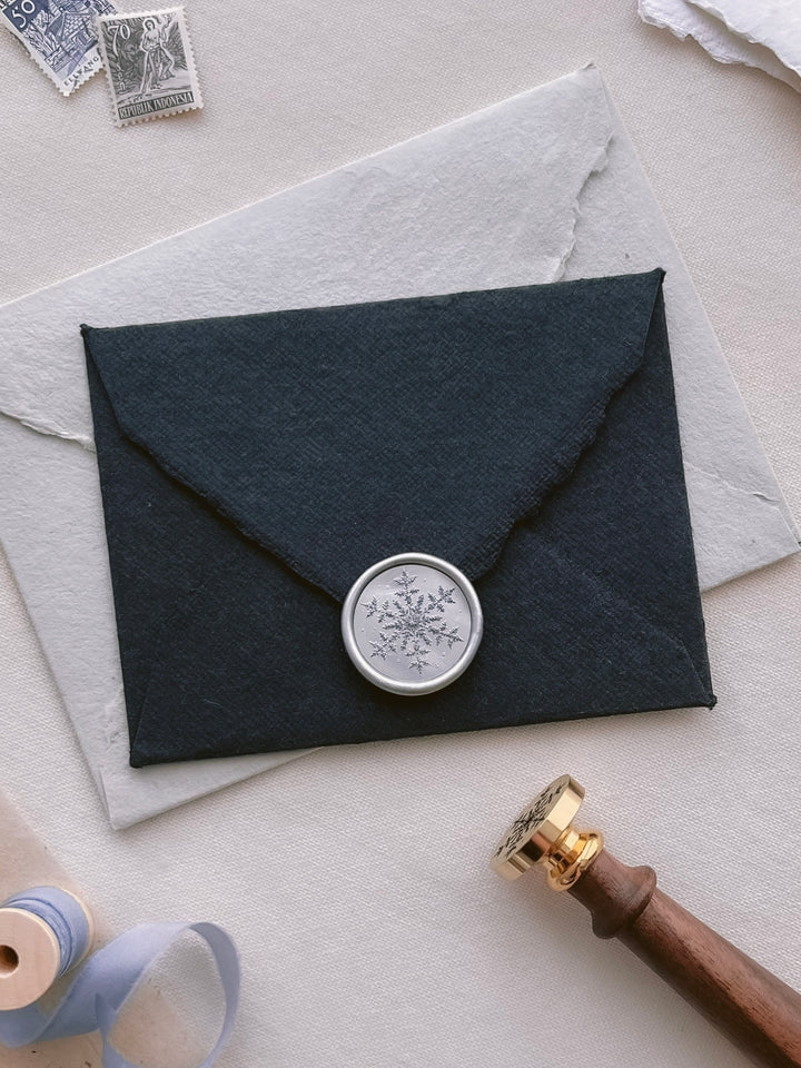 Silver snowflake wax seal with 3D engraved details on a black deckled edge handmade paper envelope next to a wax stamp head with a wooden handle