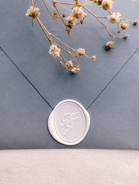 White oval monogram wax seal on a dusty blue envelope