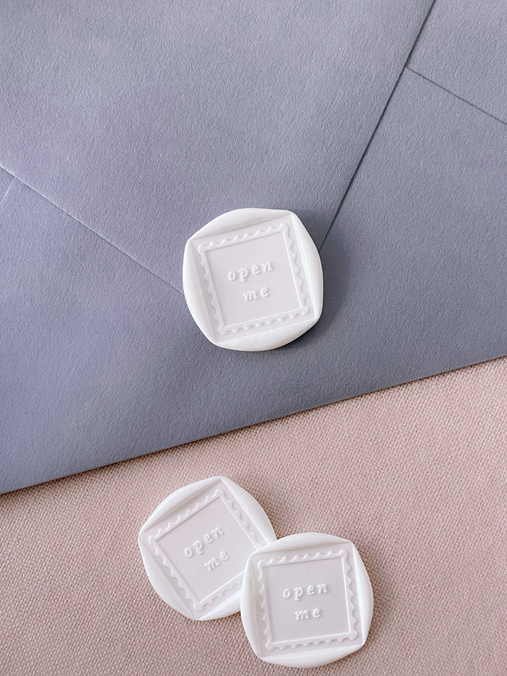 White squared shaped postage stamp style design wax seals with the phrase open me in the center