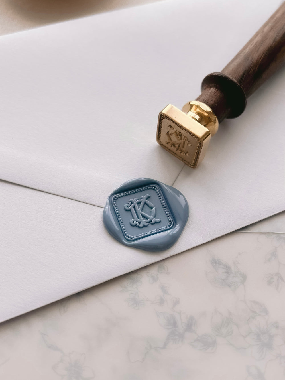 Personalized initials dusty blue square border wax seal featuring a custom vintage monogram on a white envelope with a wax stamp head on a botanical vellum jacket