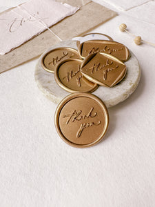 Calligraphy script "thank you" wax seals in gold and in various shapes styled with a small gray stone dish, handmade paper and a dried floral branch