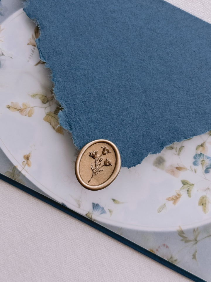 Gold tulip wax seal resting on a deckle-edge blue invitation envelope flap, set above a floral print vellum jacket over an oval wedding invitation card.