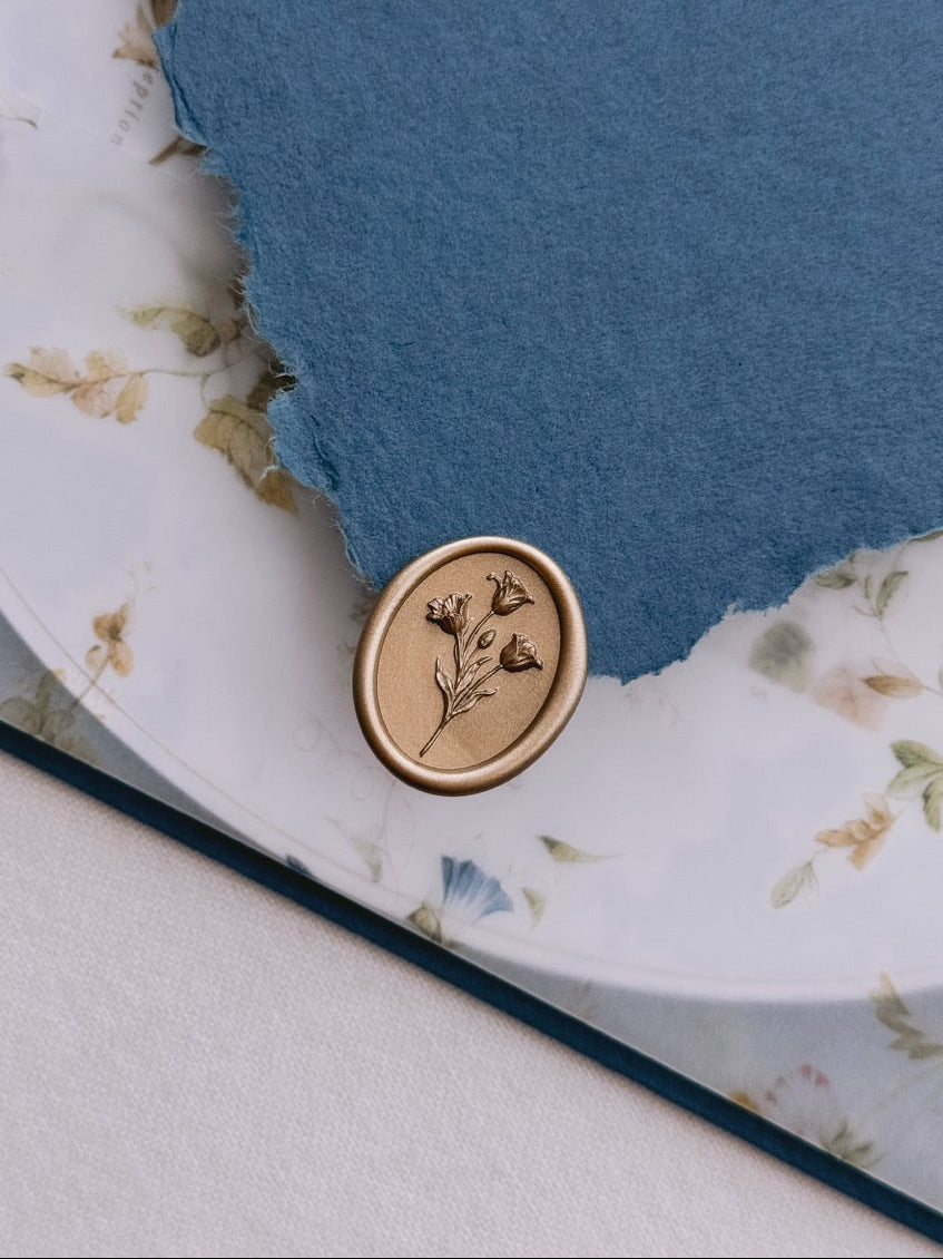 Gold tulip wax seal resting on a deckle-edge blue invitation envelope flap, set above a floral print vellum jacket over an oval wedding invitation card.