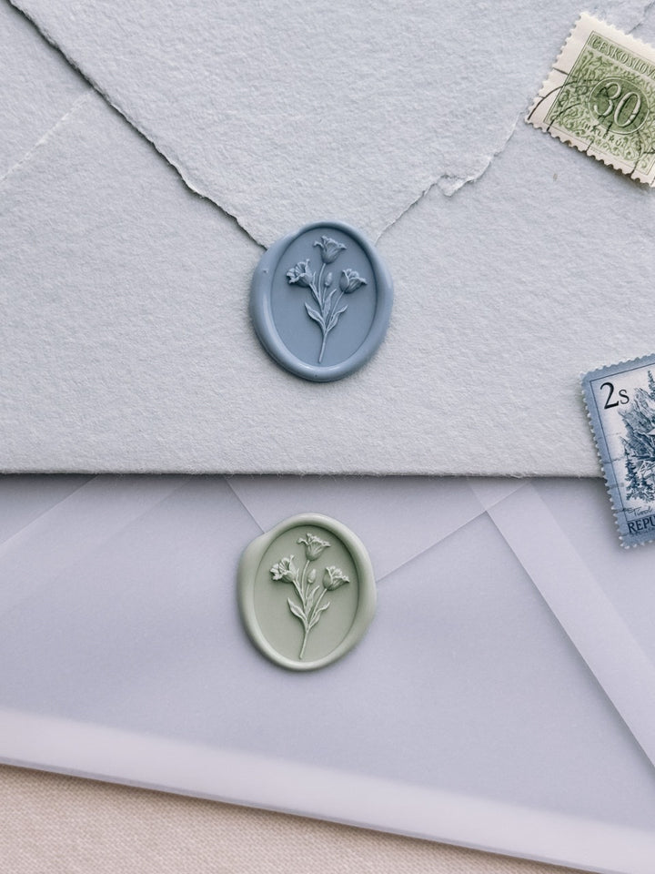 Dusty blue and sage green tulip oval wax seals on layered wedding invitation envelopes, styled with vintage postage stamps.