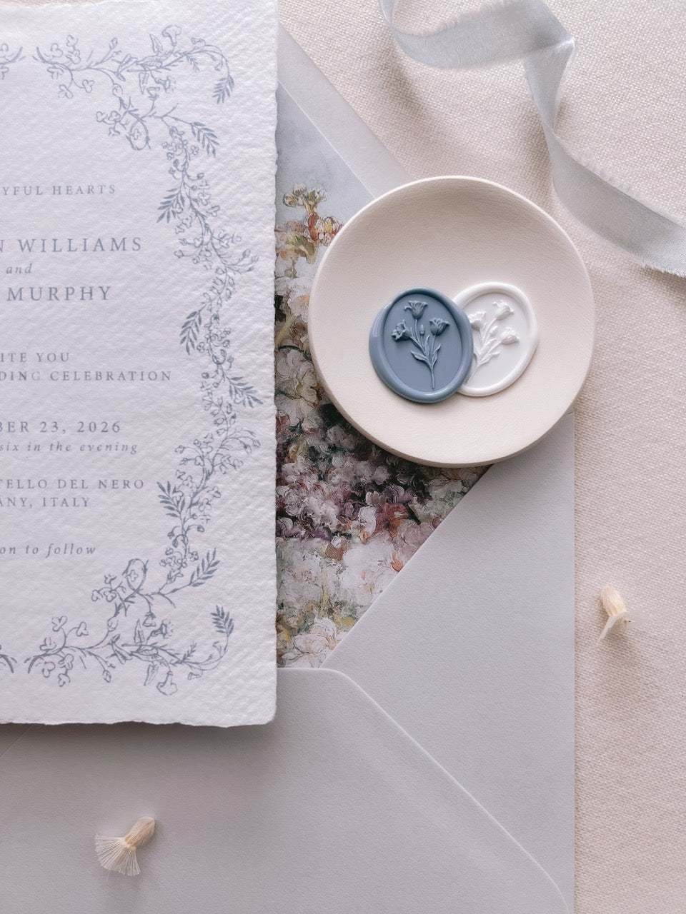 Slate blue and white tulip wax seals displayed on a ceramic dish beside a hand-illustrated botanical wedding invitation card set against a floral-lined grey envelope
