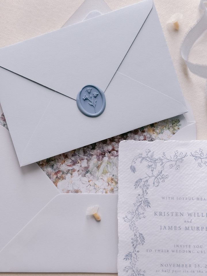Dusty blue tulip wax seal on a light blue wedding invitation envelope, layered with a floral-lined envelope and hand-torn deckle edge botanical wedding invitation card.