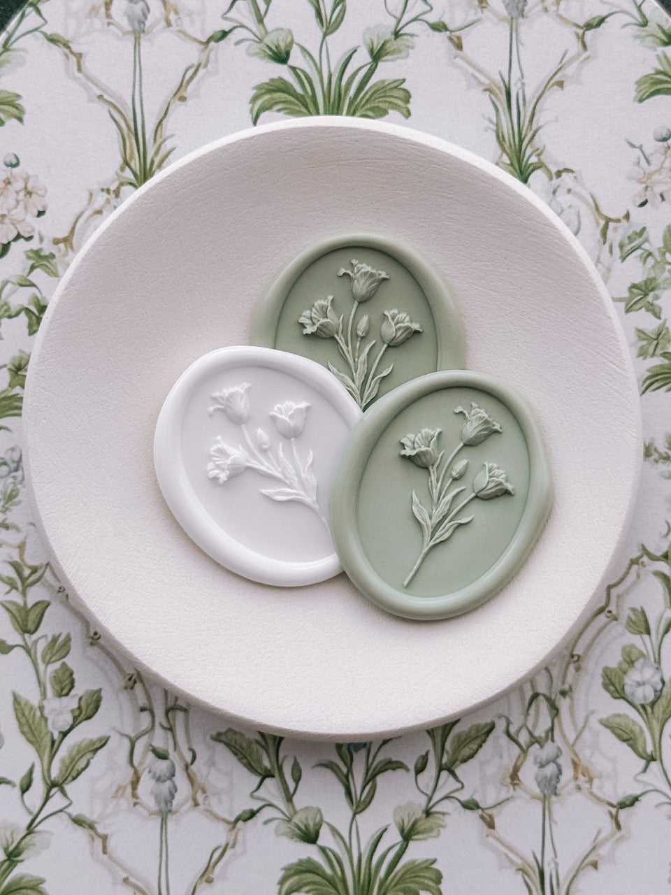Sage green and white oval tulip wax seals displayed on a ceramic dish, set against a vintage floral backdrop.