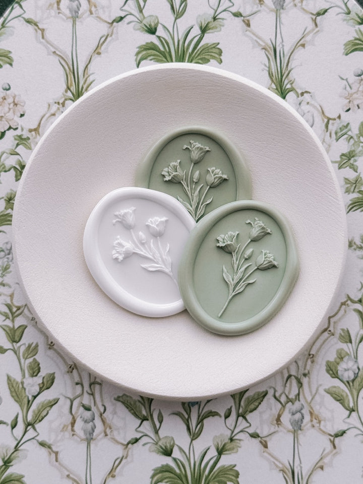 Sage green and white oval tulip wax seals displayed on a ceramic dish, set against a vintage floral backdrop.
