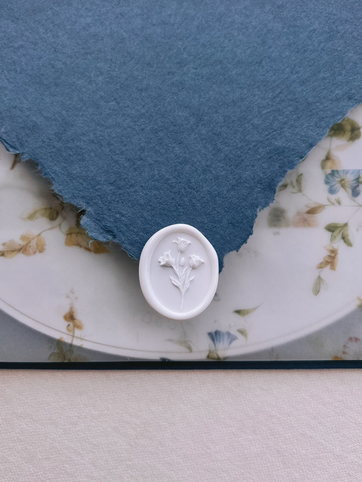 White tulip wax seal resting on a deckle-edge blue invitation envelope flap, set above a floral print vellum jacket over an oval wedding invitation card.