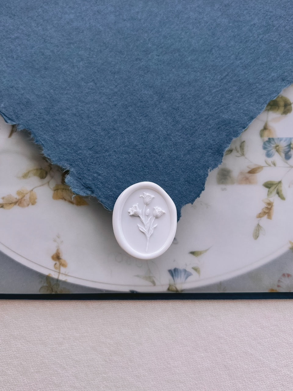White tulip wax seal resting on a deckle-edge blue invitation envelope flap, set above a floral print vellum jacket over an oval wedding invitation card.