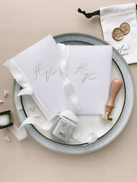 A set of two His and Her gold foil vellum vow books in calligraphy script with soft white colored silk ribbon styled with bride wedding rings, wax seal stamp, spool of soft white silk ribbon, gold wax seals and white linen pouch