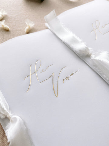 A set of two His and Her gold foil vellum vow books in calligraphy script with soft white colored silk ribbon
