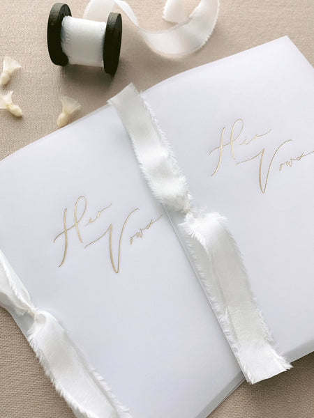 A set of two His and Her gold foil vellum vow books in calligraphy script with soft white colored silk ribbon styled with spool of soft white silk ribbon