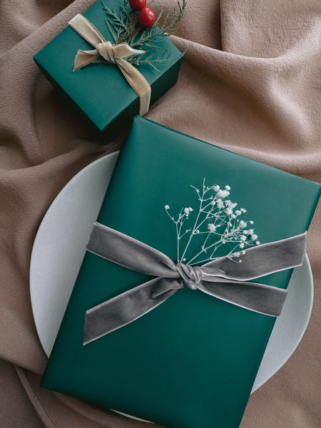 green Christmas gift wrap decorated with silver velvet ribbon bow and a dried floral branch and another gift with gold velvet ribbon and leaf branch