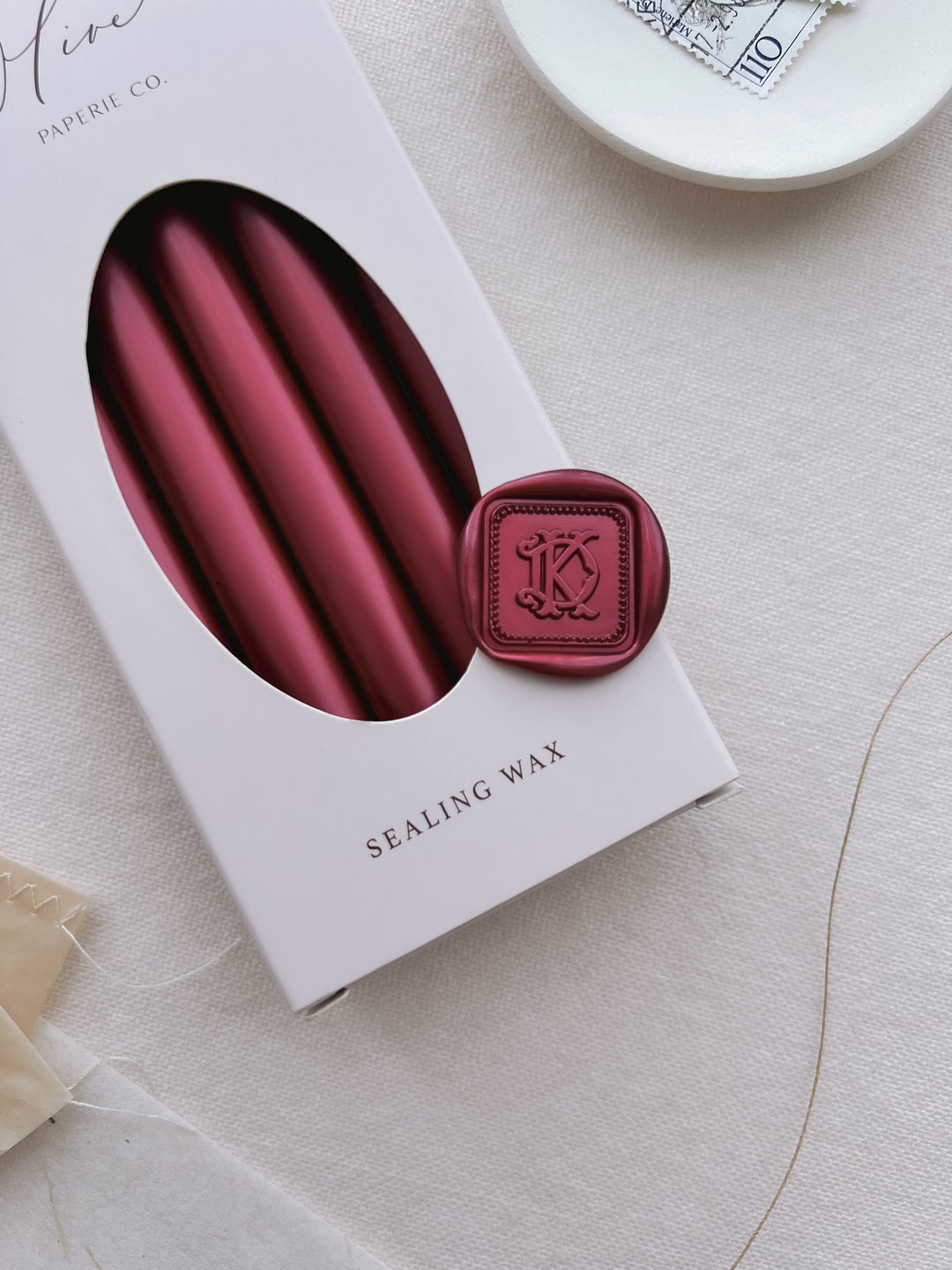 Dark metallic red personalized initials vintage square border wax seal featuring a custom monogram on a box of dark metallic red sealing wax sticks on a light beige background
