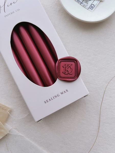 Dark metallic red vintage interlocked monogram square shaped wax seal with a border design, on a box of metallic red sealing wax sticks