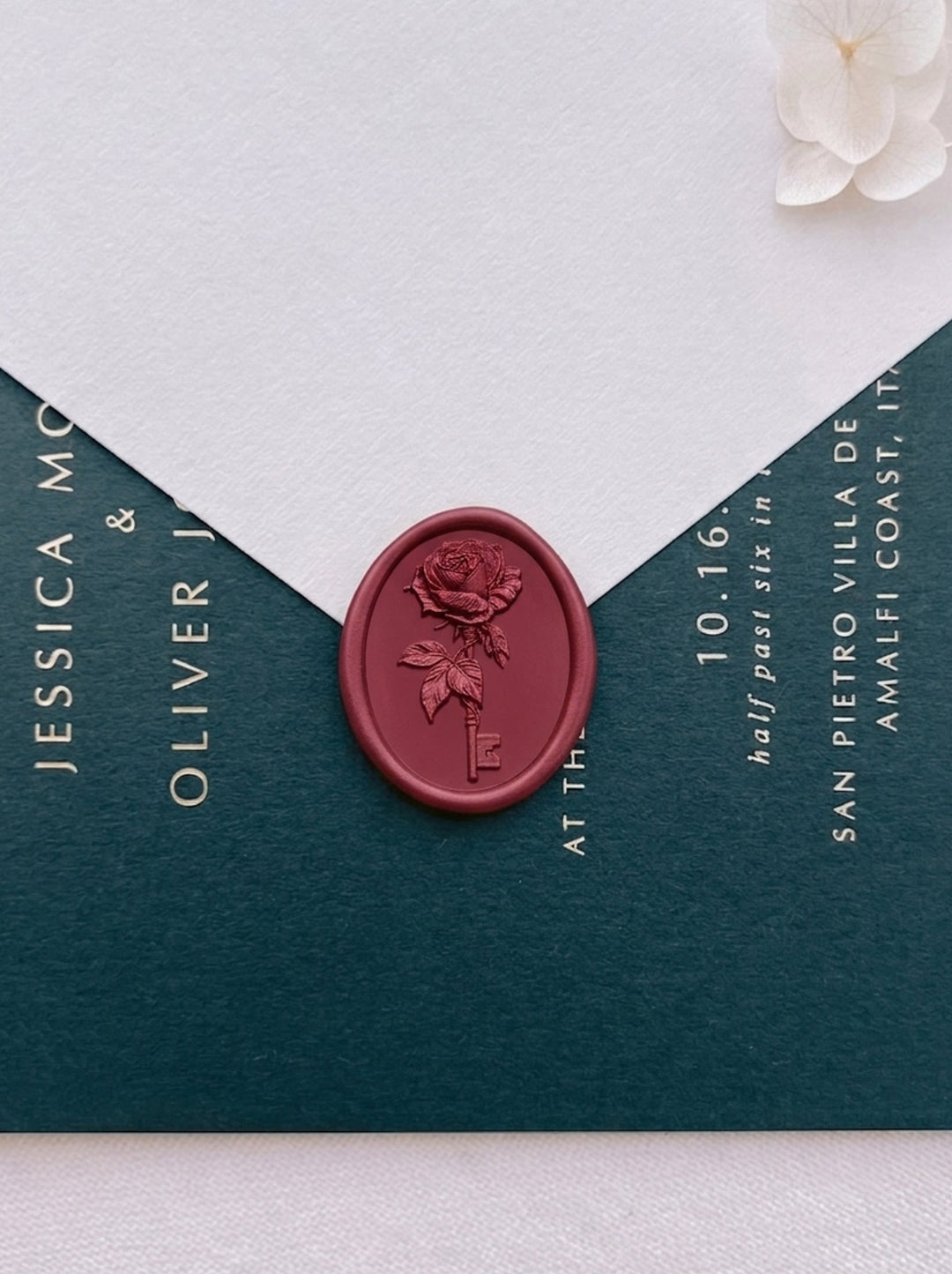 Dark metallic red ornate rose key oval wax seal on a beige envelope flap over a dark green gold foil wedding invitation card