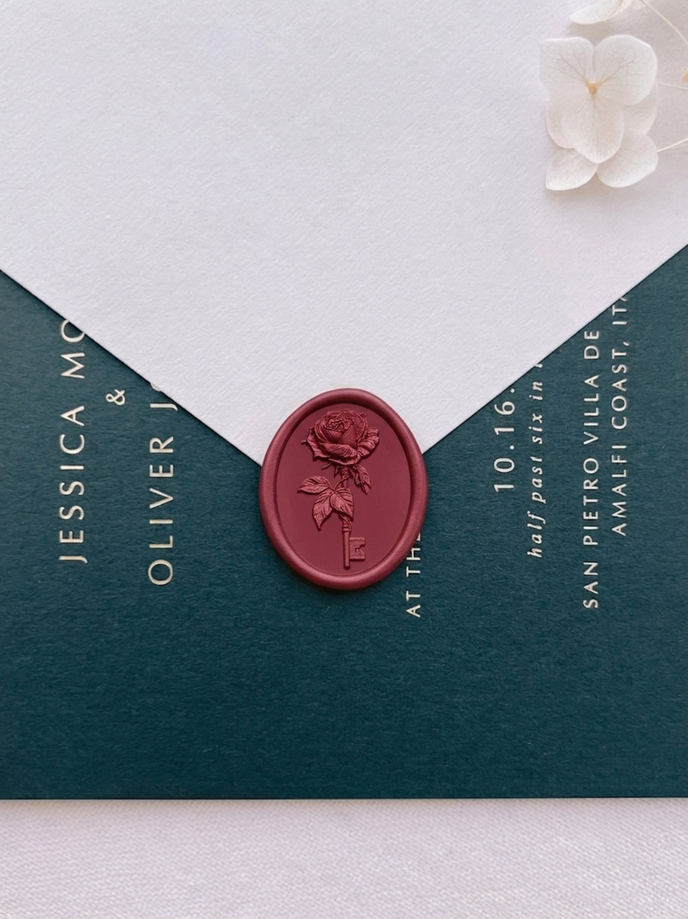 Dark metallic red rose and key wax seal on a white wedding envelope, sealed against a deep green wedding invitation card with gold foil typography.