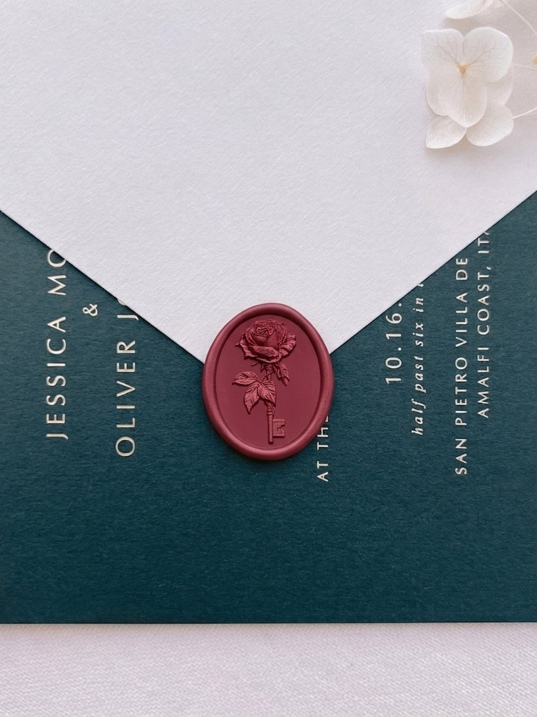 Dark metallic red rose and key wax seal on a white wedding envelope, sealed against a deep green wedding invitation card with gold foil typography.