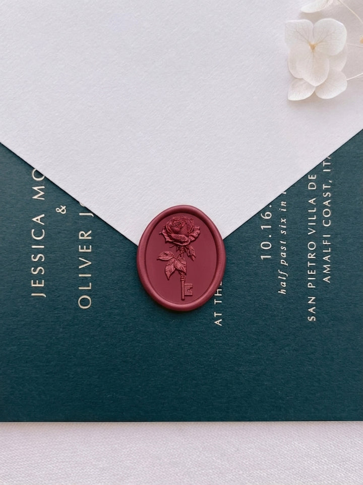 Dark metallic red rose and key wax seal on a white wedding envelope, sealed against a deep green wedding invitation card with gold foil typography.