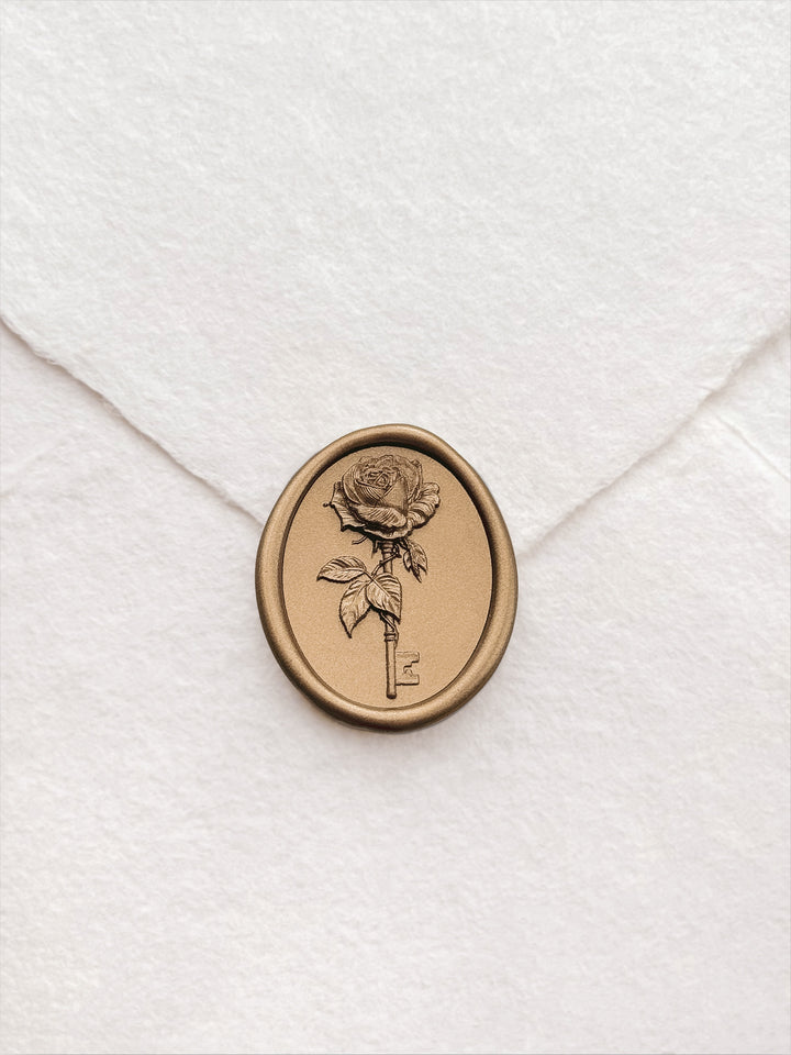 Gold rose and key wax seal on a deckle-edge ivory wedding envelope, close-up showing intricate 3D engraving of a full bloom rose and skeleton key motif.