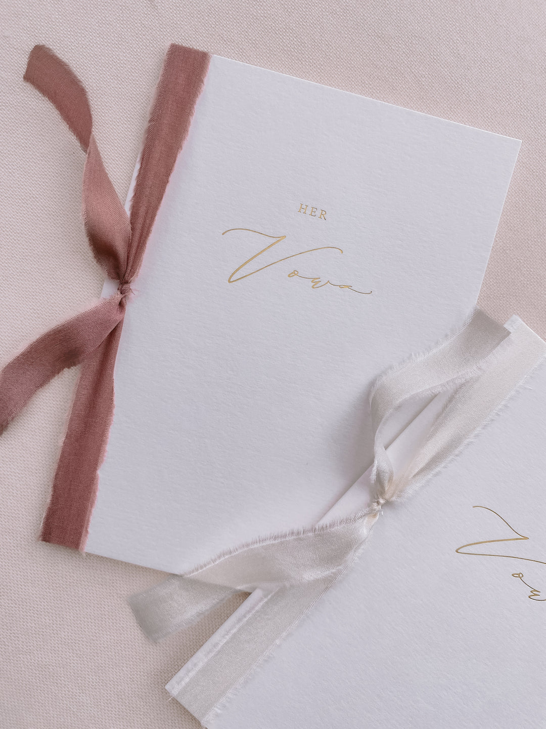 A set of two His and Her gold foil white vow books in calligraphy script with one tied in dusty rose colored silk ribbon and the other in soft white colored silk ribbon