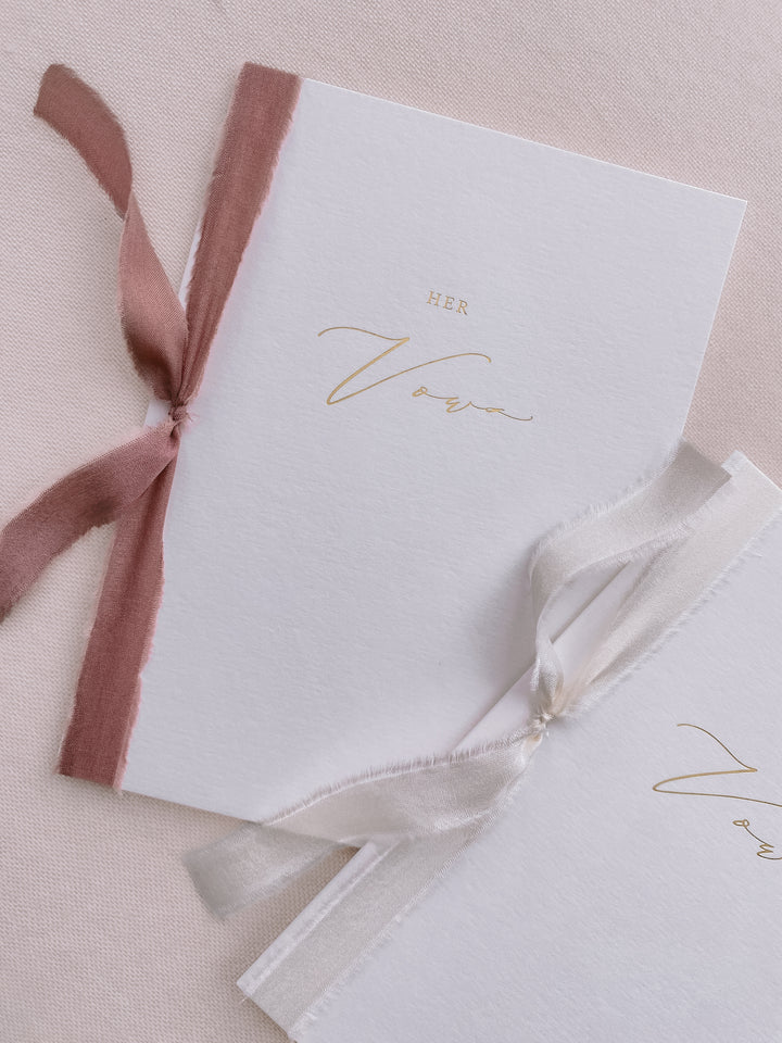 A set of two His and Her gold foil white vow books in calligraphy script with one tied in dusty rose colored silk ribbon and the other in soft white colored silk ribbon