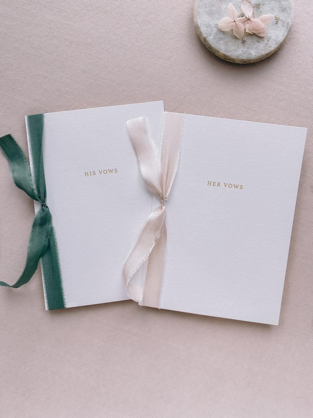 A set of two His and Her gold foil white card stock vow books in typeface font with one tied in antique white colored silk ribbon and the other in forest green colored silk ribbon