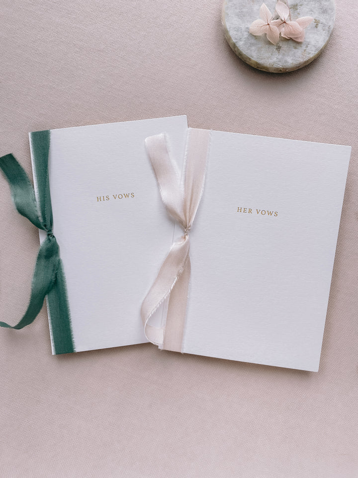 A set of two His and Her gold foil white card stock vow books in typeface font with one tied in antique white colored silk ribbon and the other in forest green colored silk ribbon