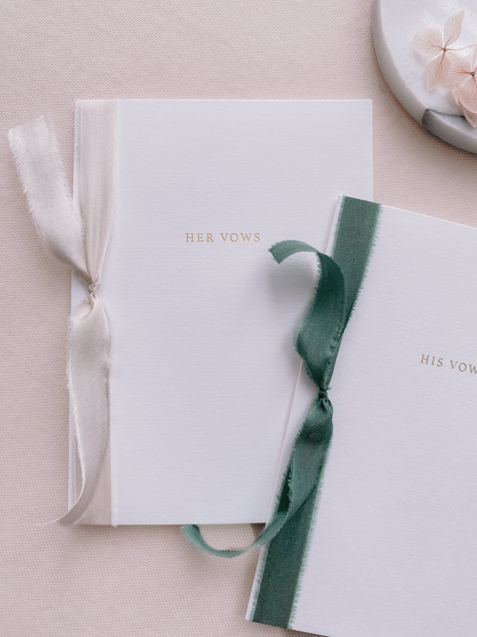 A set of two His and Her gold foil white card stock vow books in typeface font with one tied in antique white colored silk ribbon and the other in forest green colored silk ribbon