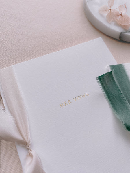 A set of two His and Her gold foil white card stock vow books in typeface font with one tied in antique white colored silk ribbon and the other in forest green colored silk ribbon